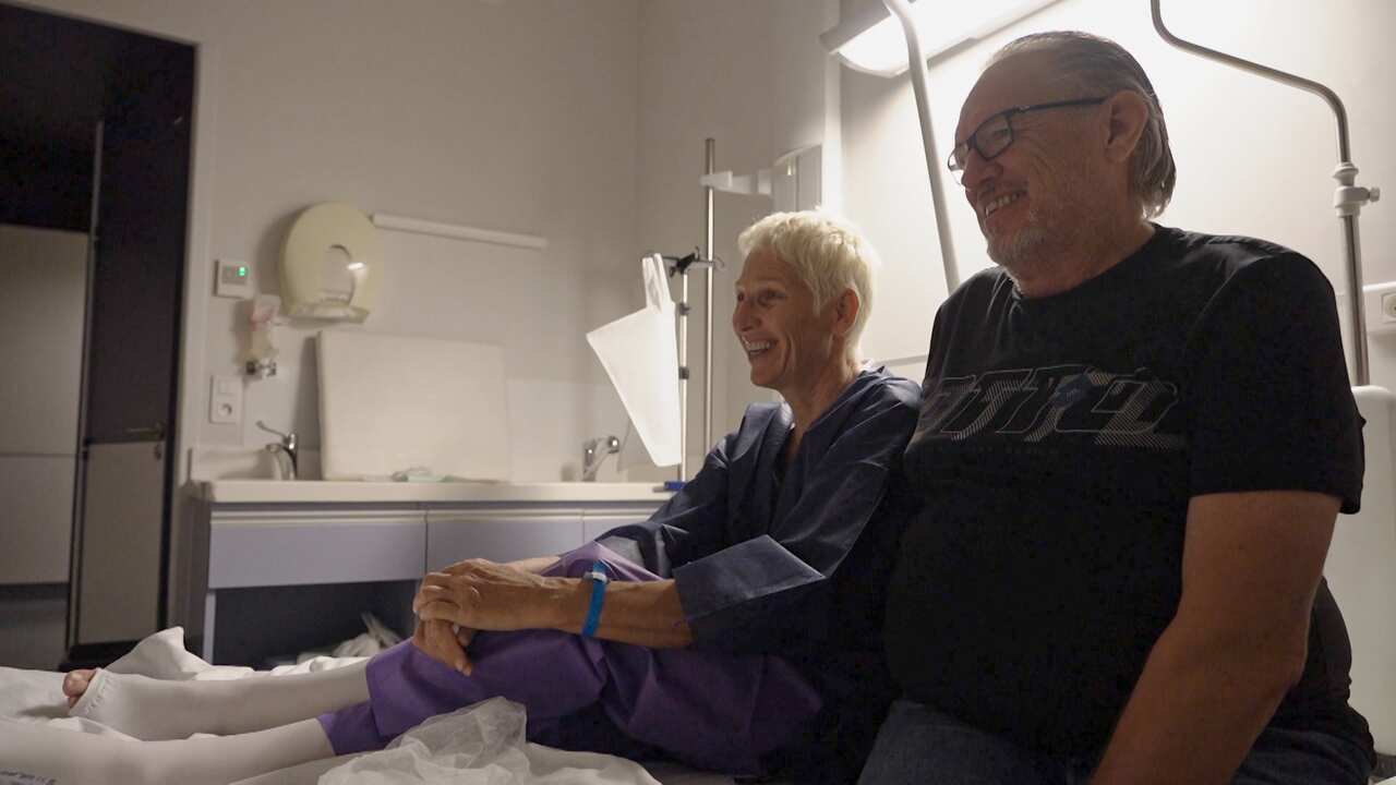 A woman and a man. The woman is sitting in a hospital bed and the man is sitting beside her