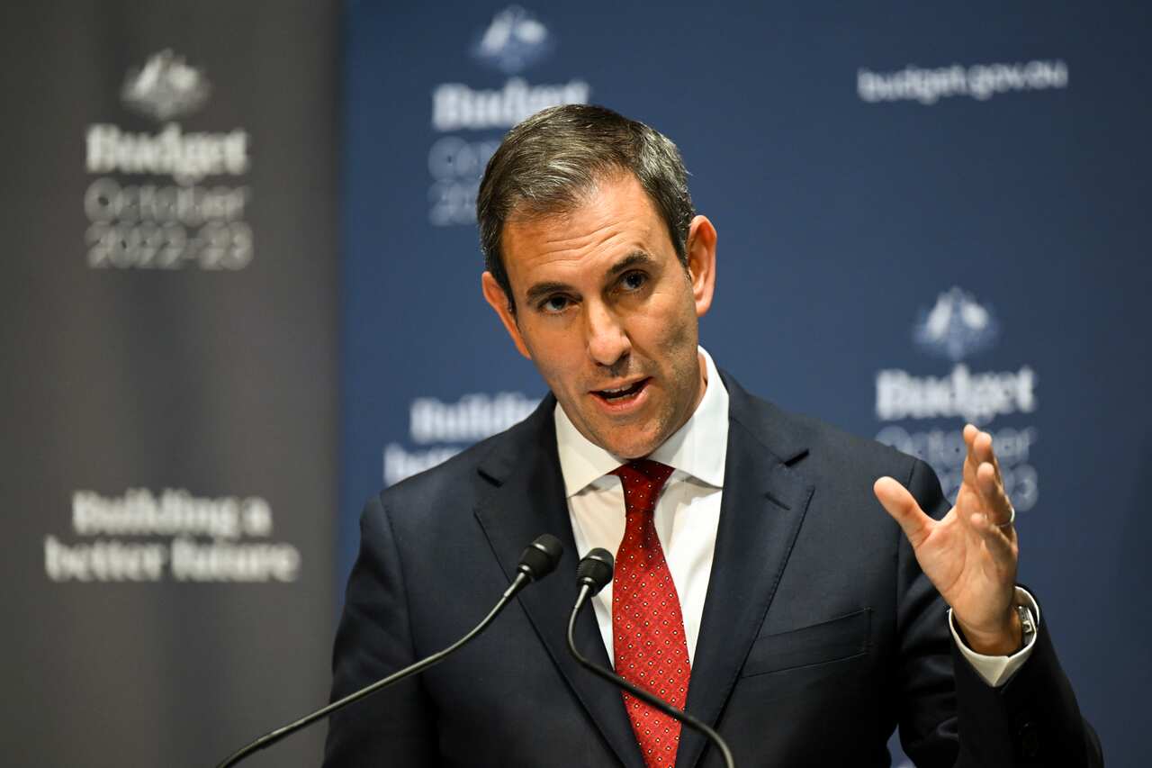 A man wearing a dark suit and red tie gestures as he speaks into a microphone