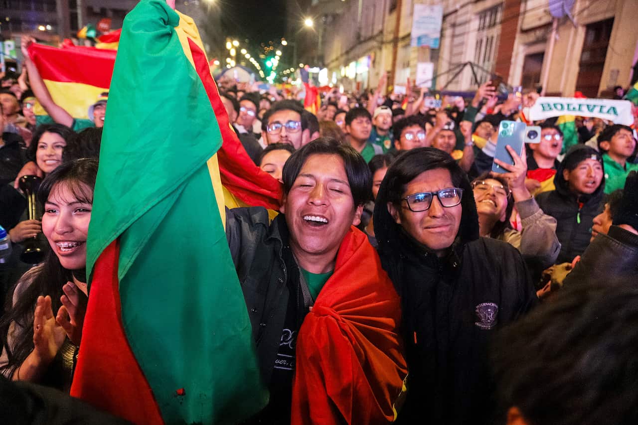A jubilant crowd with faces of excitement and joy celebrate on a street at night, waving Bolivian flags and banners
