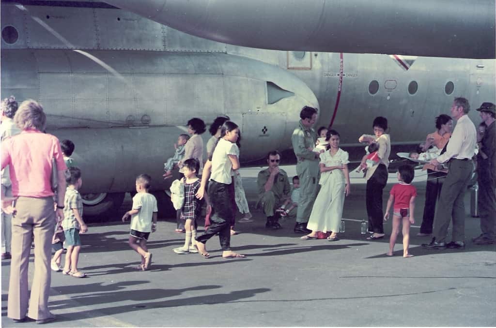 Around a dozen babies and children, and a dozen adults standing and sitting on the tarmac beside a large grey RAAF military aircraft. The adults looking after the children.