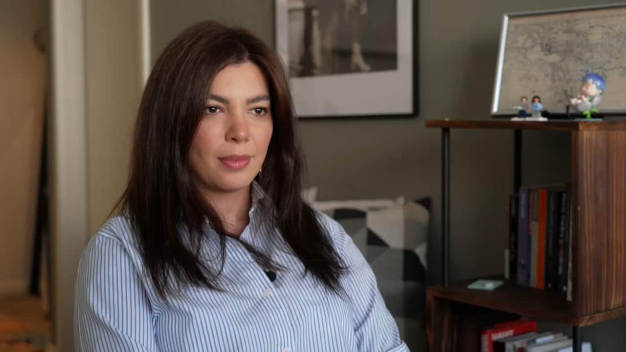 A woman with long dark hair wearing a striped shirt sits indoors, with a bookshelf and framed picture in the background.