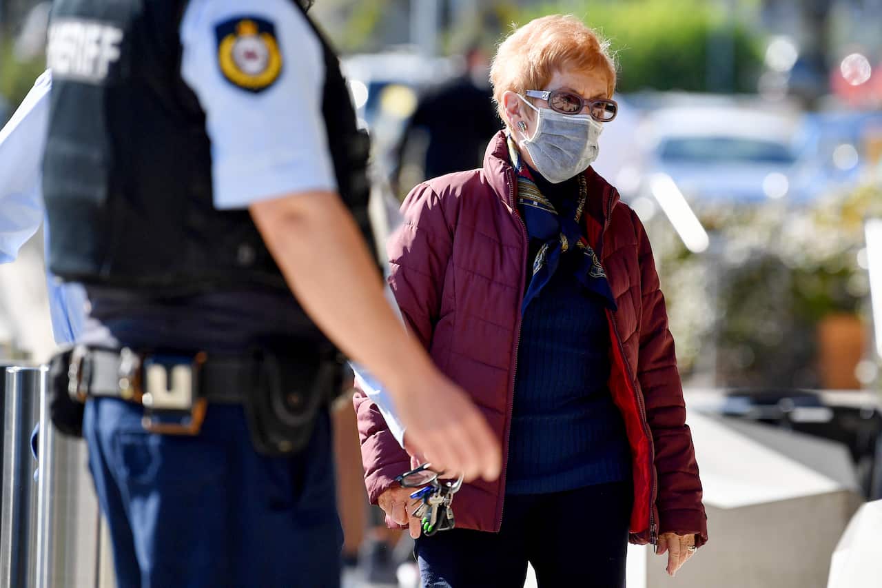 An elderly woman wearing sunglasses and a face mask walks past a police officer