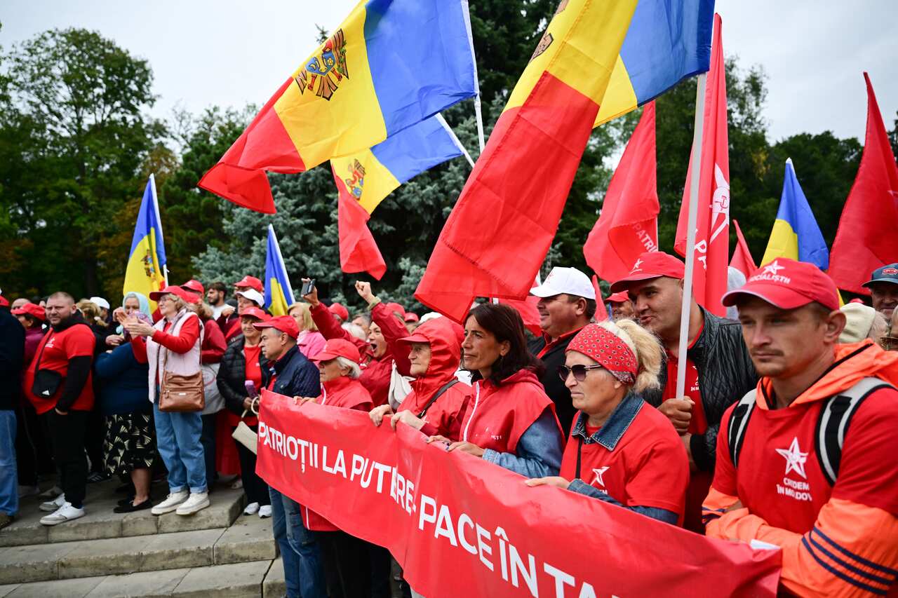 A group of people dressed in red outfits and carrying red, yellow and blue flags walk behind a red banner with white writing on it on a road. There are also people standing on the side of the road