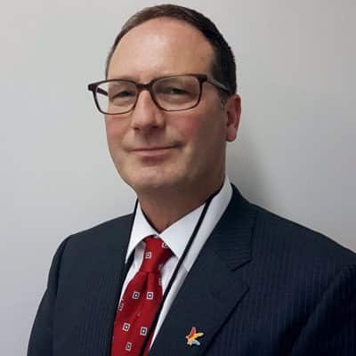 A white man with brown hair weaing brown framed glasses, a navy pinstriped blazer, white shirt, and red tie.