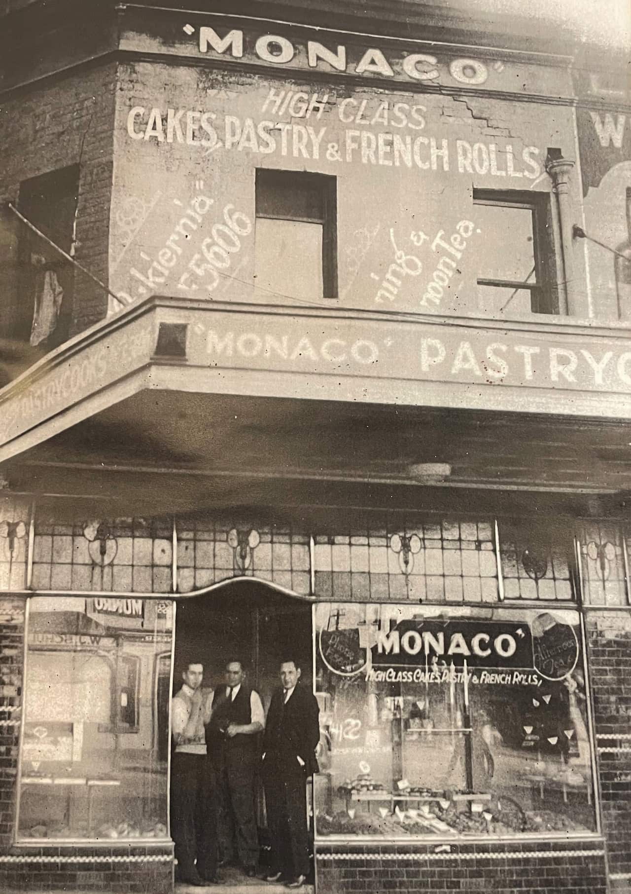 A black and white photograph of a cake shop with staff standing in the doorway.