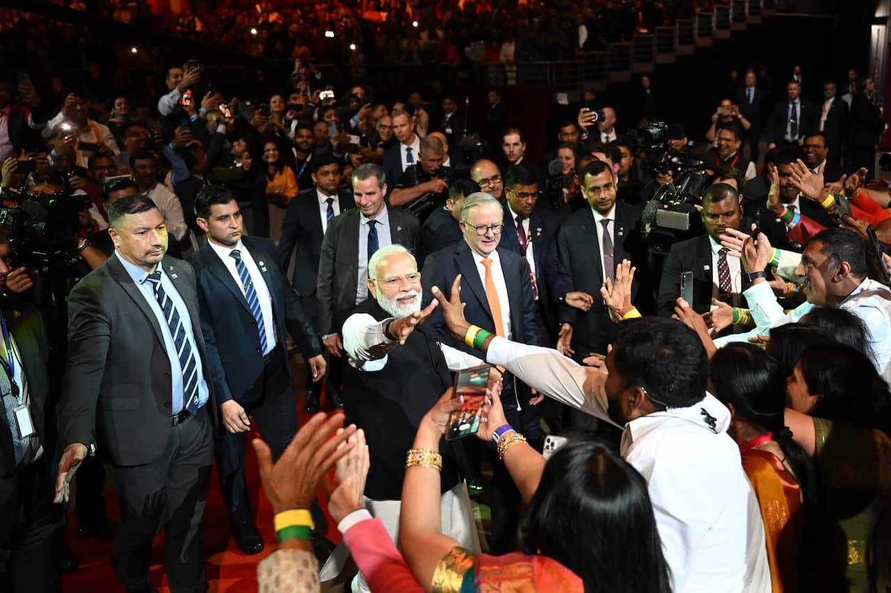 Anthony Albanese (right) and Narendra Modi stand among a large crowd, with attendees reaching out to shake Modi's hand, as both leaders are flanked by security personnel.