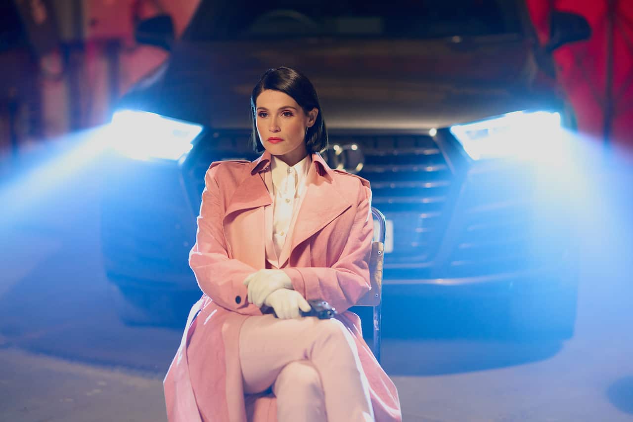 A woman is an elegant pants-and-shirt set with long pinkish coat sits on a chair in front of a car. She holds a gun in one gloved hand.