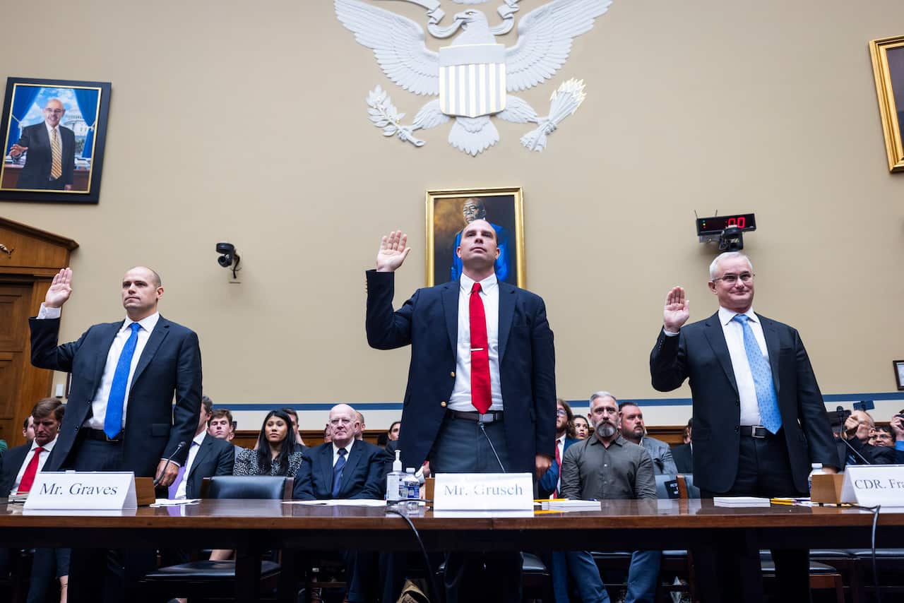 Three men wearing suits and ties standing in front of a desk. Each have one hand in the air.