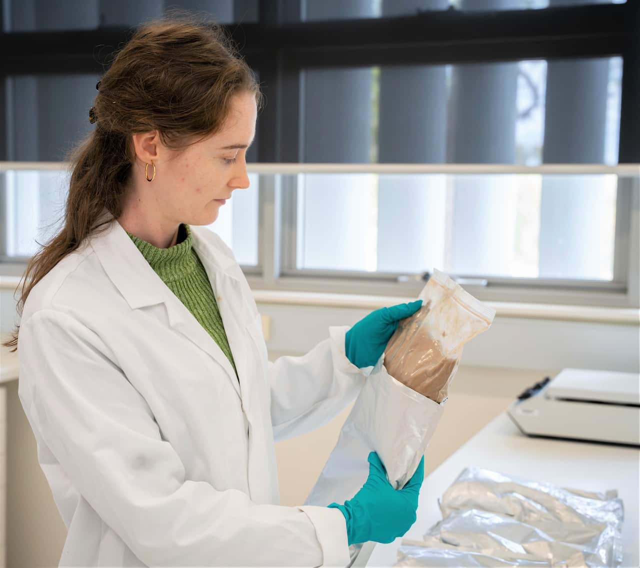 A woman with brown hair wearing a white lab coat holds a plastic packet of brown powder.