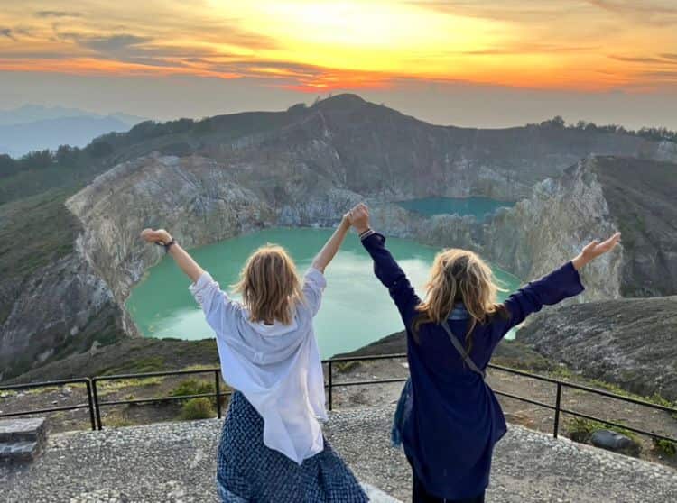 Sharon and Rosa Carol, seen from behind, standing in front of the basin of Indonesia's Kelimutu volcano at sunrise, throwing their hands in the air.