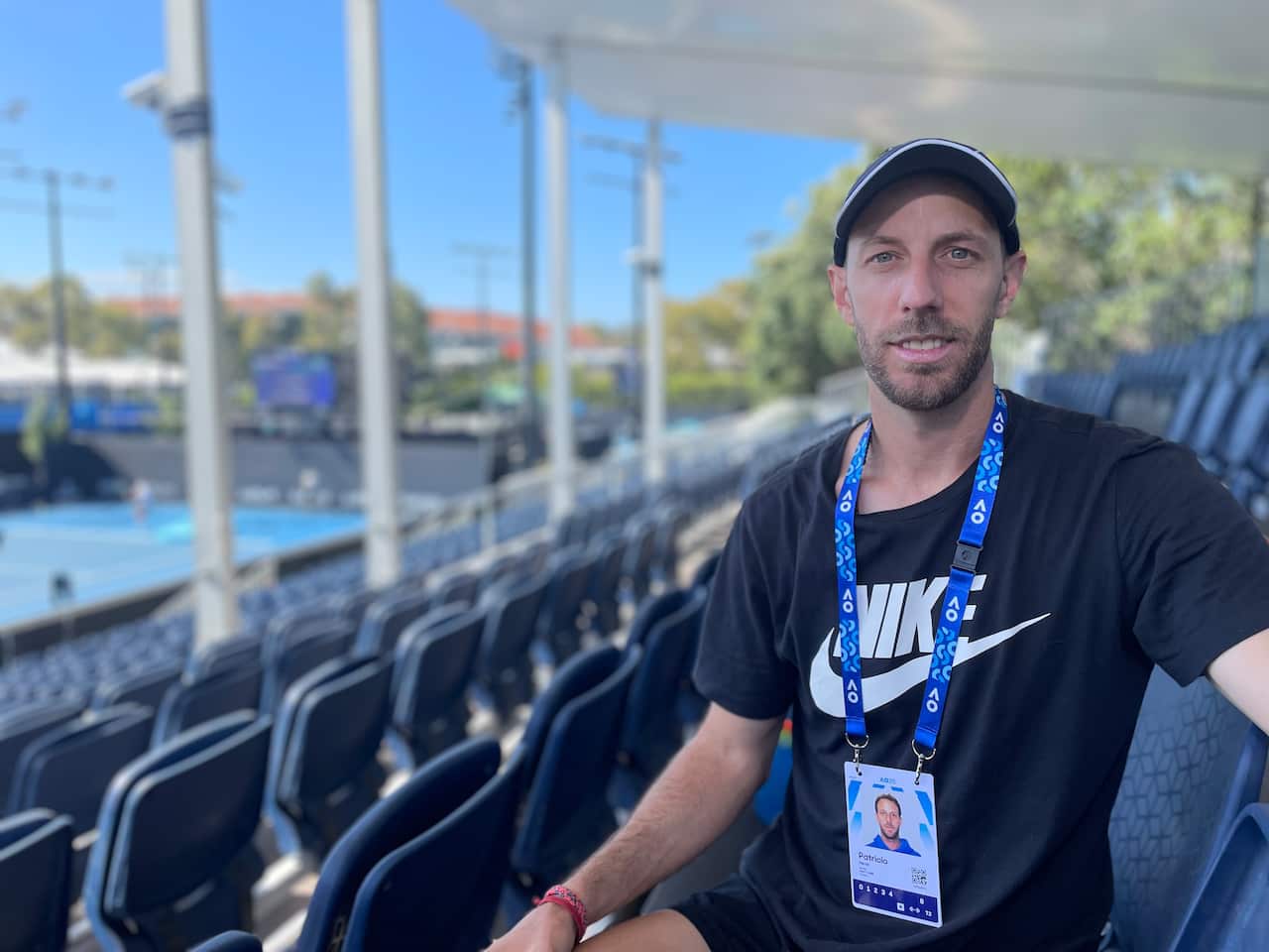 A man wearing a black Nike T-shirt and a cap and a blue lanyard smiles while sitting in a tennis stand.