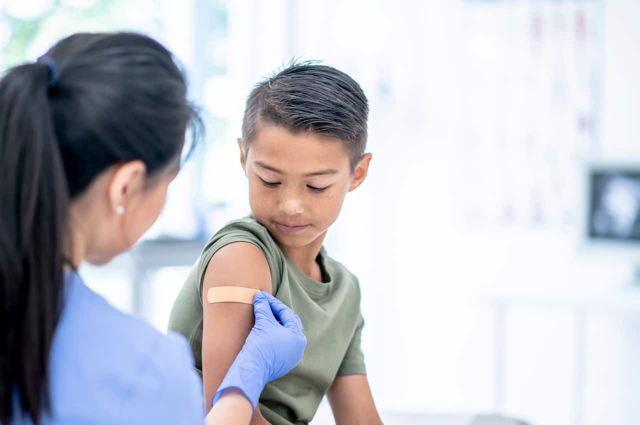 A doctor wearing blue scrubs and blue gloves places a band-aid on a young boy's arm.