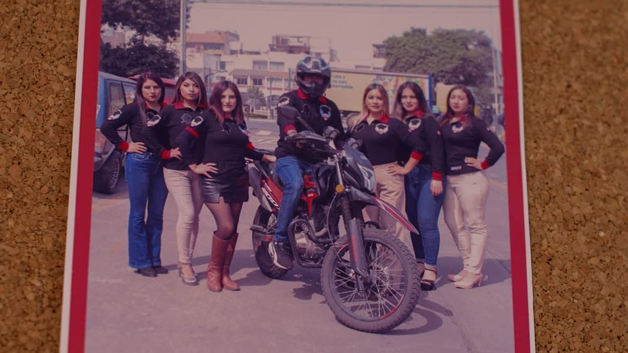 A photo of a photograph showing a group of women in matching black and red long-sleeve t-shirts standing with their hands on their hips, on either side of a man in a matching shirt riding a motorbike. 