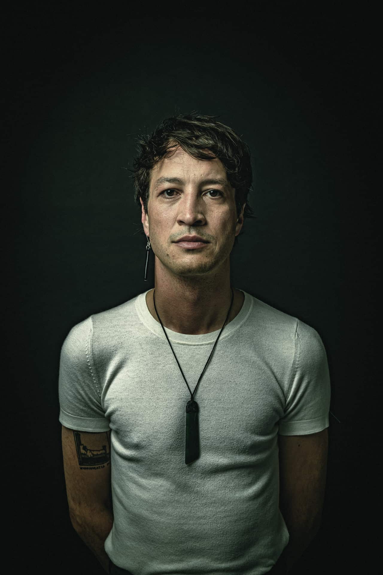 A man in a white shirt against a black background. He is wearing a long pendant around his neck and a long earring in his right ear.