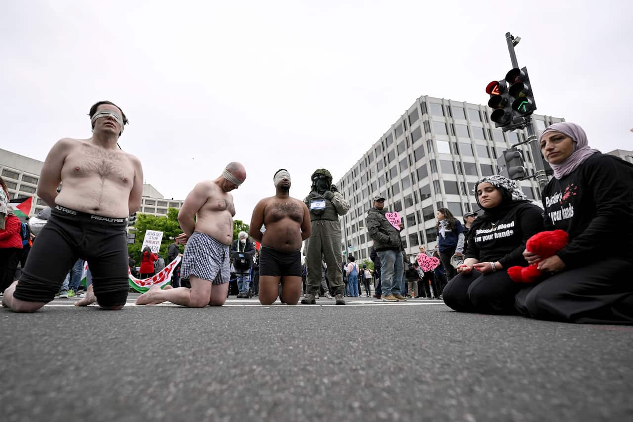 Several men wearing only underpants kneel on a road, next to a group of women and other protesters.