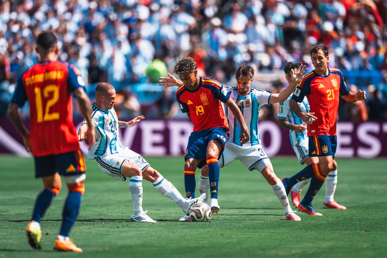 A footballer wearing a red shirt and blue shorts fights for possession of the ball against multiple defenders.