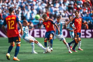 A footballer wearing a red shirt and blue shorts fights for possession of the ball against multiple defenders.