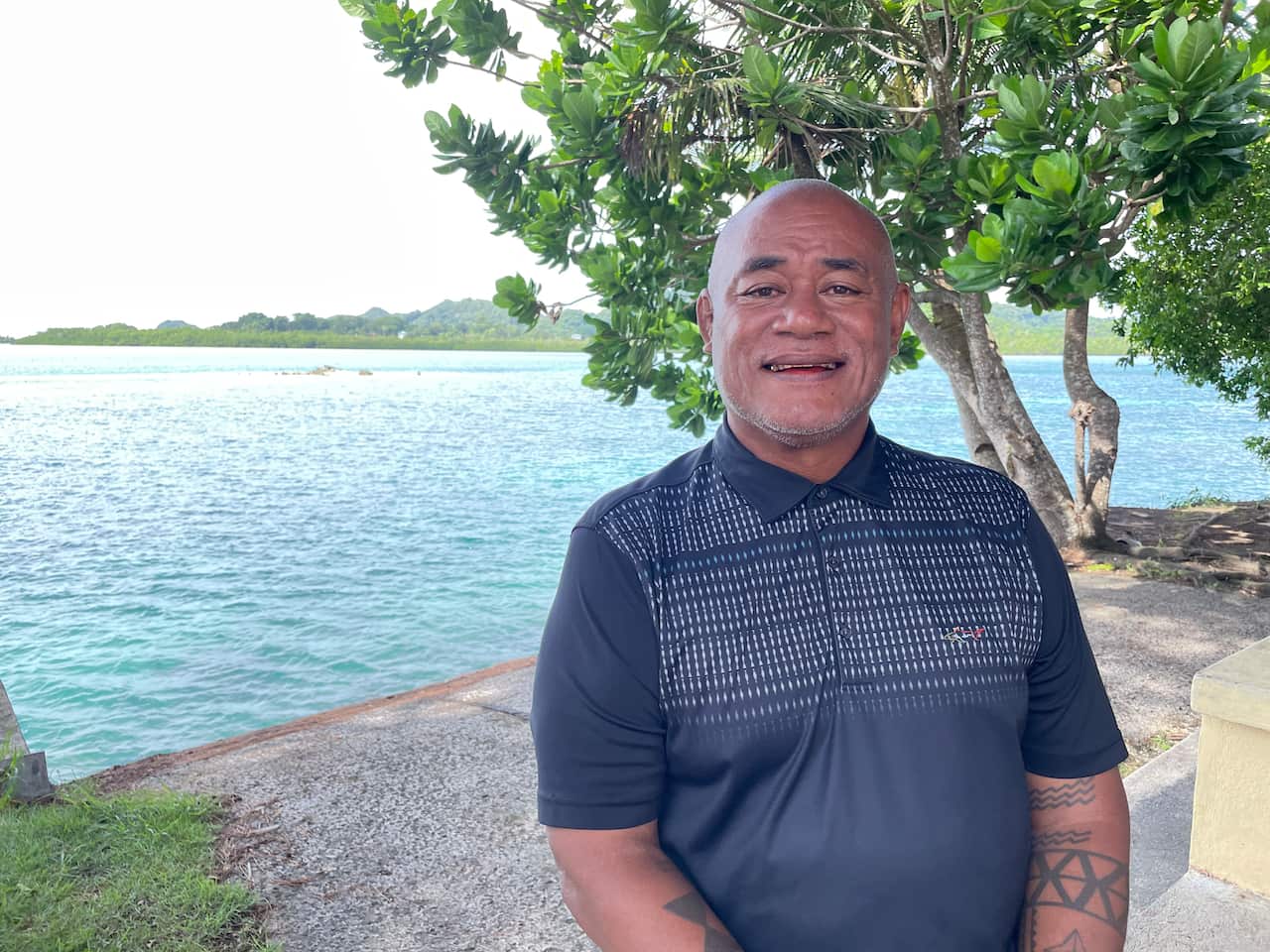 An elderly man in a blue T-shirt smiles as he stands in front of the blue lagoon.
