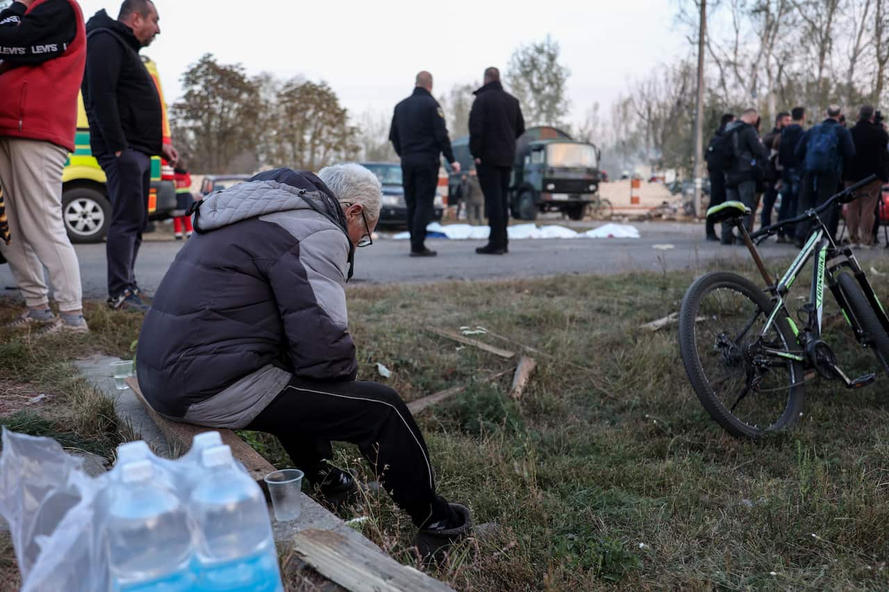Man in grey and navy blue puffer jacket and tracksuit pants sits on a plank of wood with his head hung low. Emergency crews gather in the distance behind him.