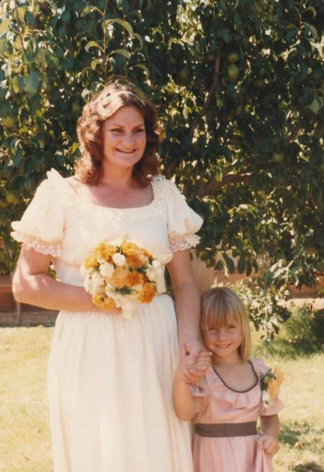 A woman in a white dress smiles as she holds flowers in one hand and the hand of a little girl in the other, who is also smiling.
