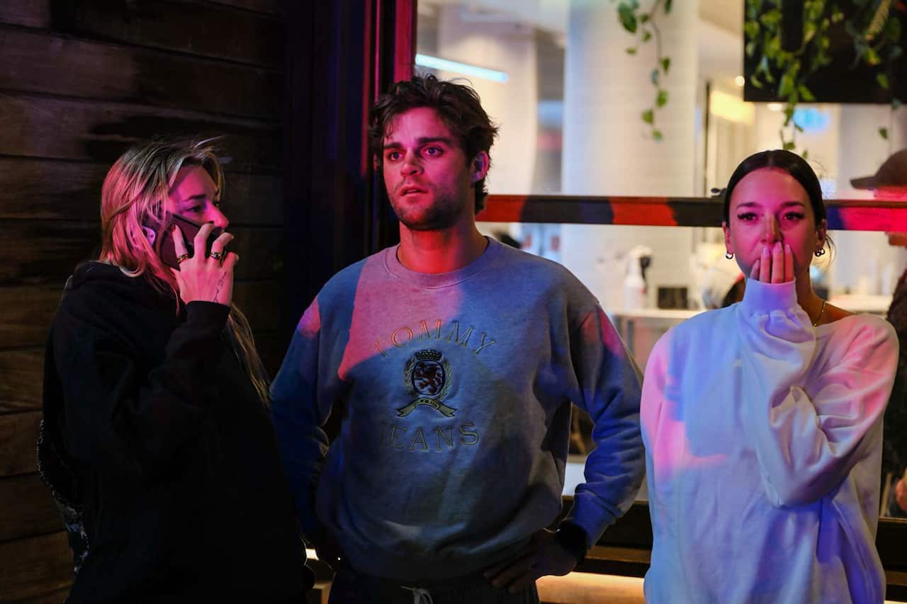 Three people stand outside at night, looking distressed, with coloured emergency lights reflecting on them.