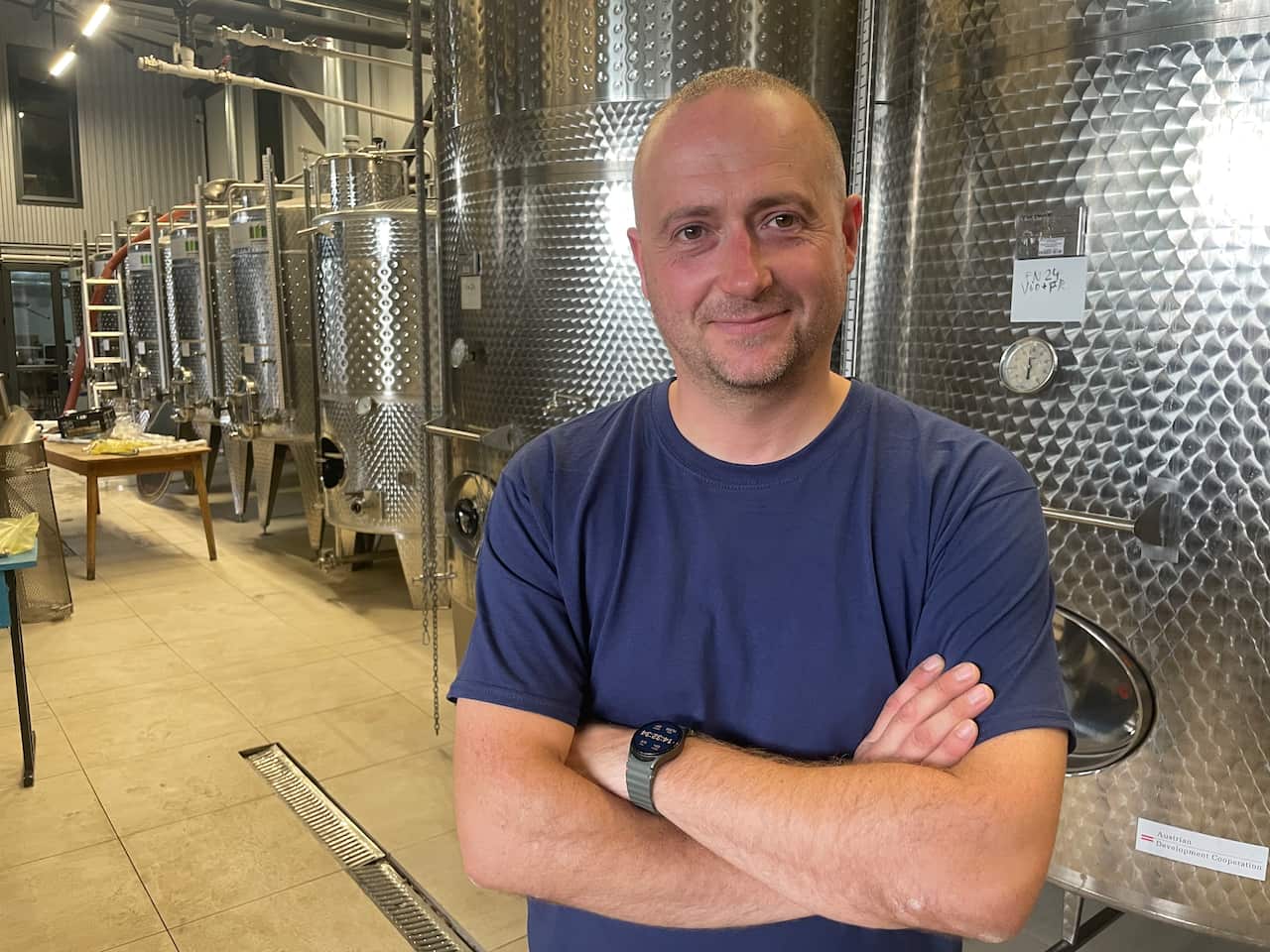 A middle aged man wearing a navy t-shirt with his arms crossed in front of large silver tanks used for making wine. 