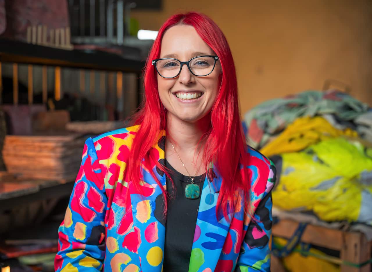 A woman with pink hair and a multi-coloured jacket sits in a warehouse.