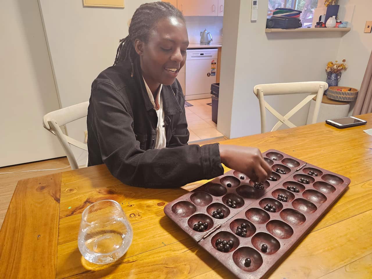 Chantal wears a dark jacket and plays with a brown igisoro board and marbles at a wooden table 