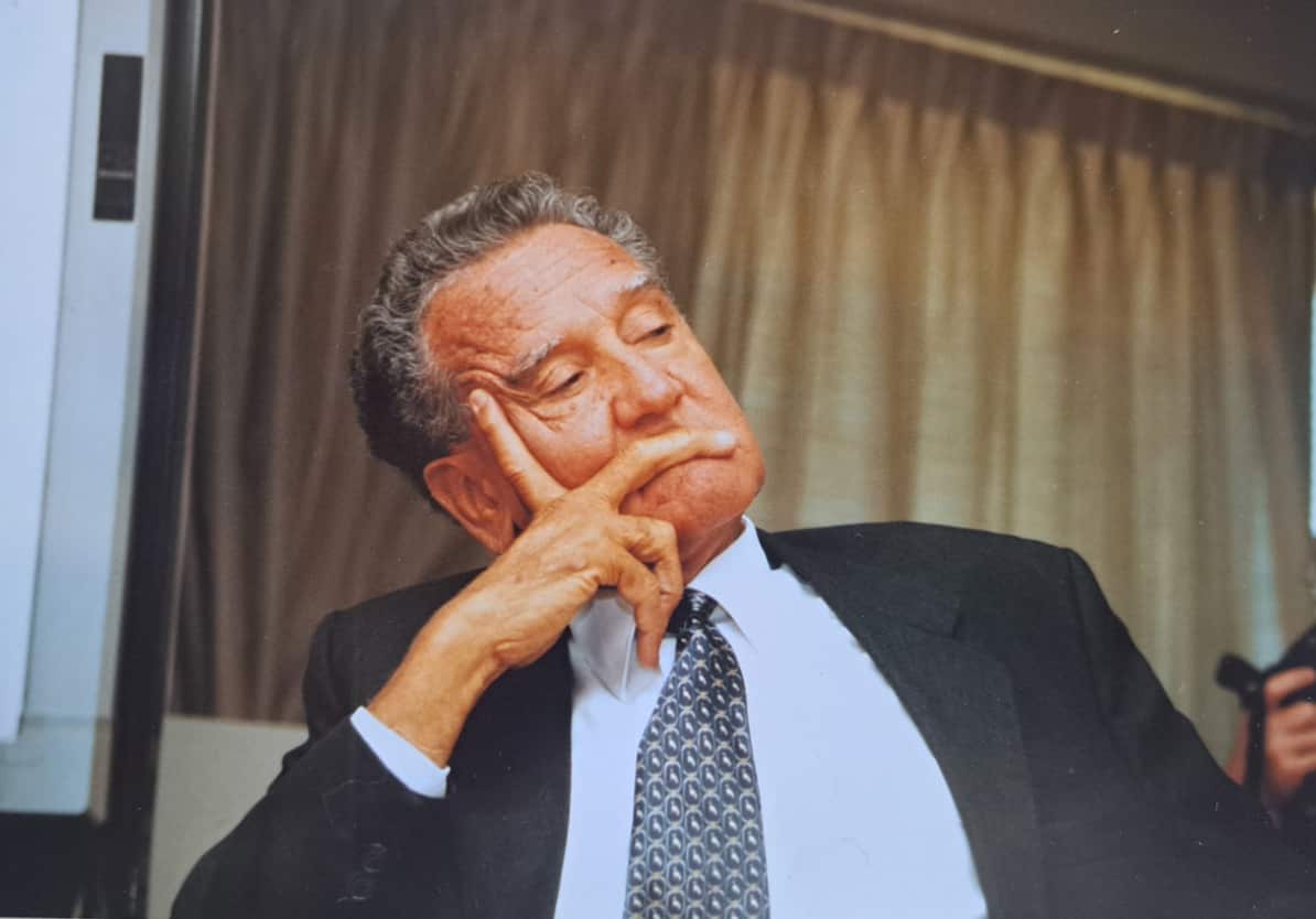 Charles Perkins sitting down, wearing a black suit, light blue shirt and tie.