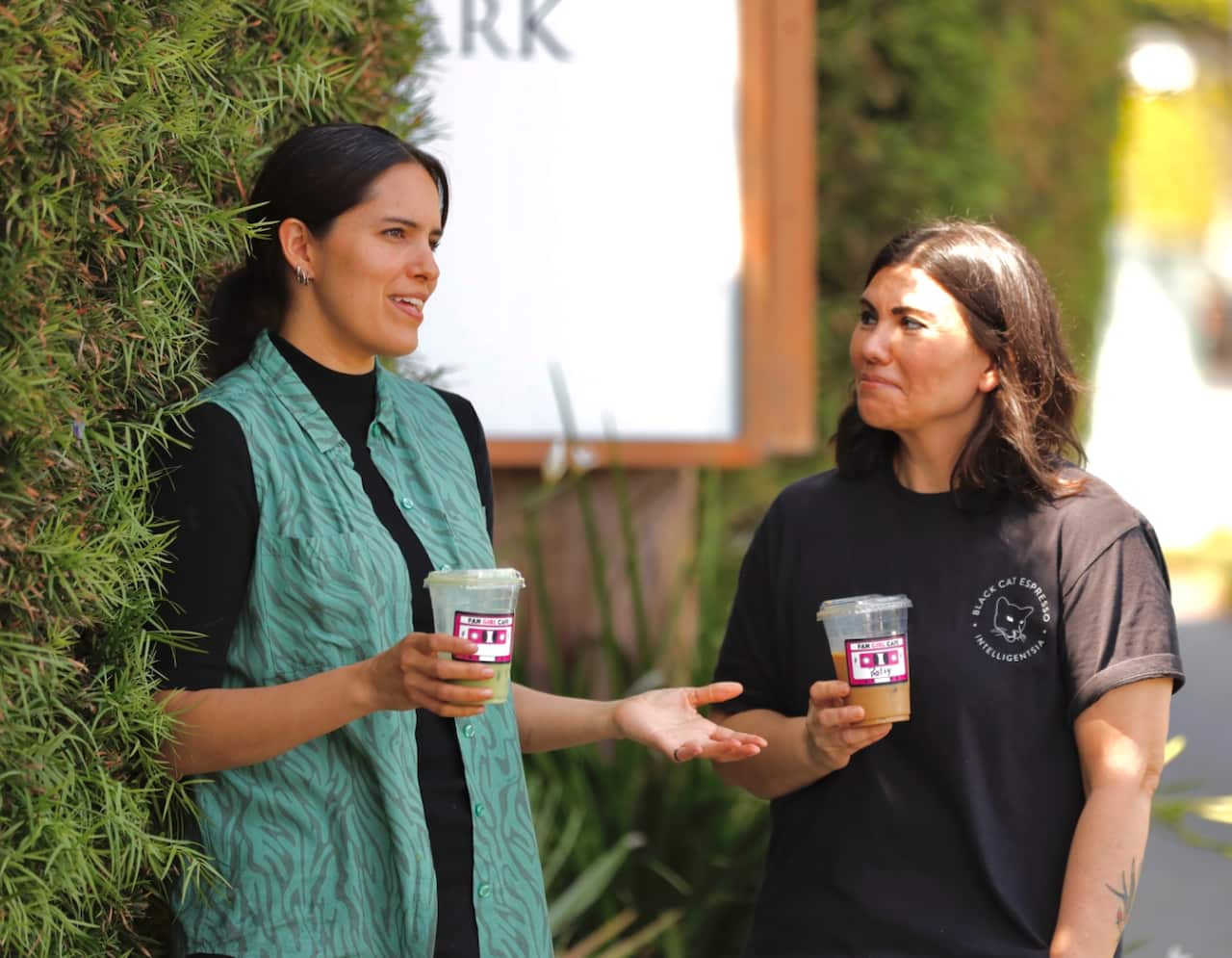 Two women holding plastic coffee cups are talking to each other as they stand outside.