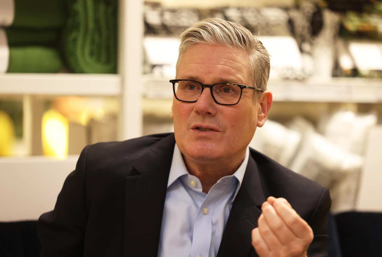 Keir Starmer, wearing a black jacket and blue shirt, waving his hand slightly.