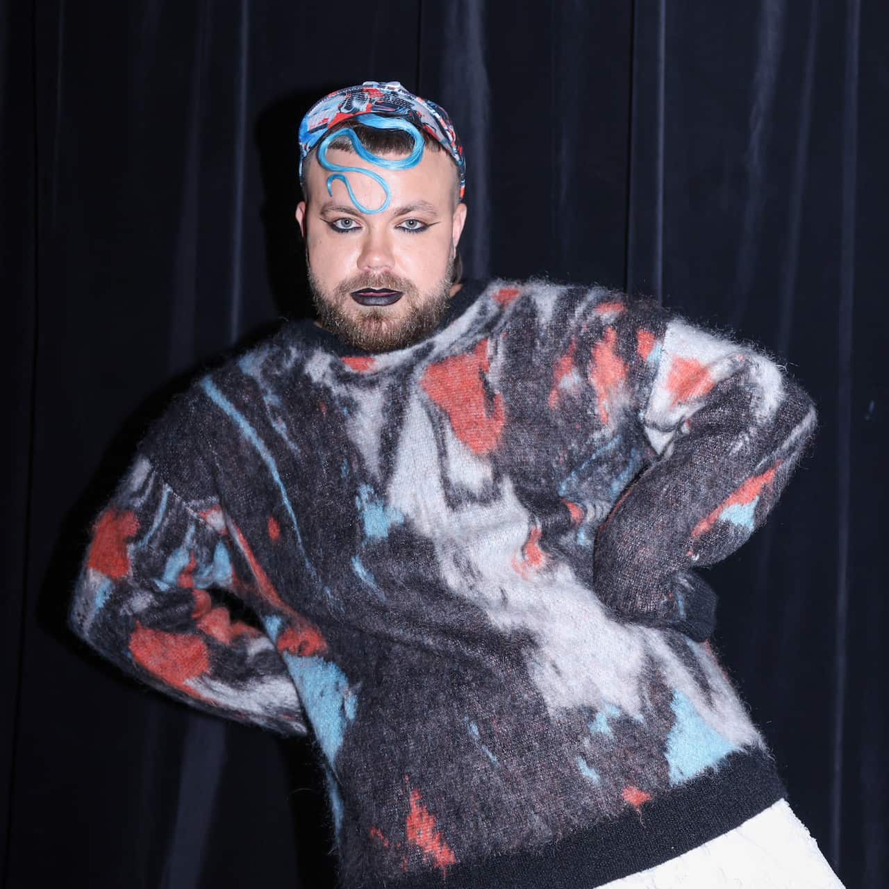 A person in a jumper and head scarf with a grey, black, red and blue pattern. The model has a beard and is wearing black lipstick