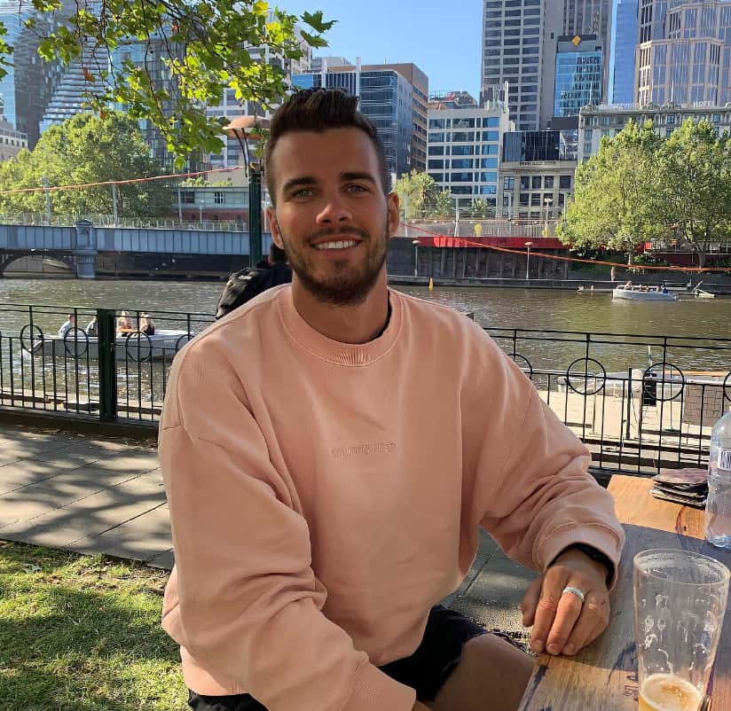 A man wearing a peach-coloured jumper, sitting at a bench next to a river