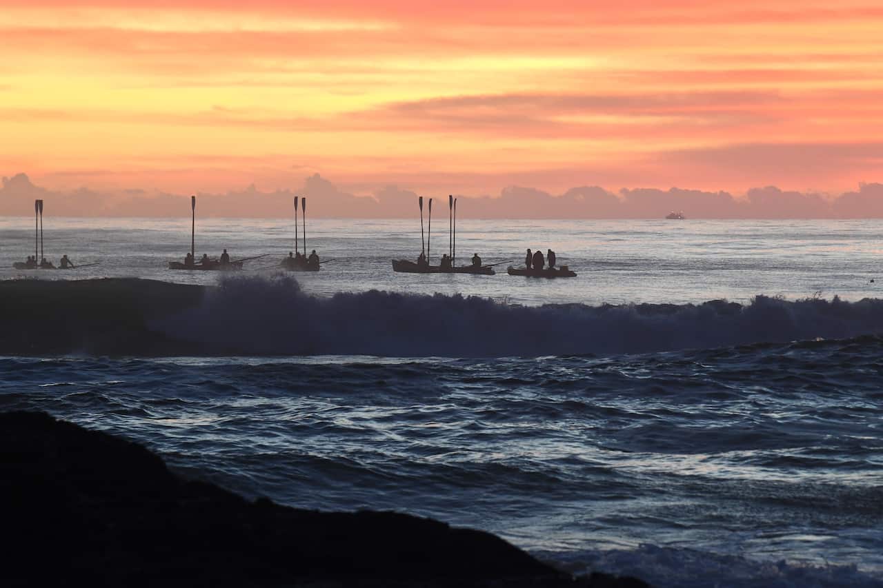A small huddle of surf boats, with paddles raised, silhouetted against the dawn sky while small waves break in the foreground.