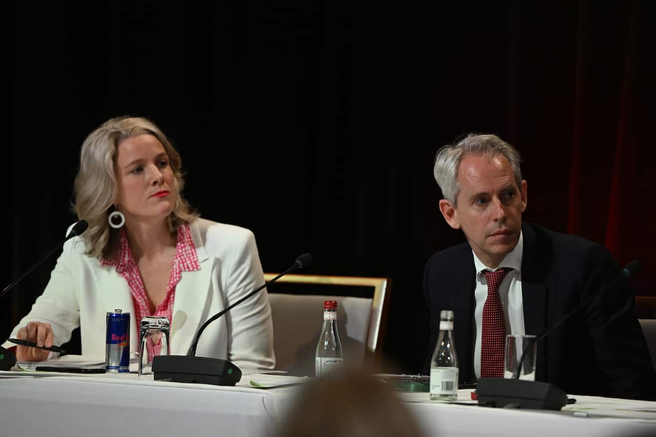 A woman in a white jacket sits at a conference table with a man in a blue suit