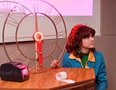 A young woman in a blue dress with yellow lapels sits next to a red science experiment.