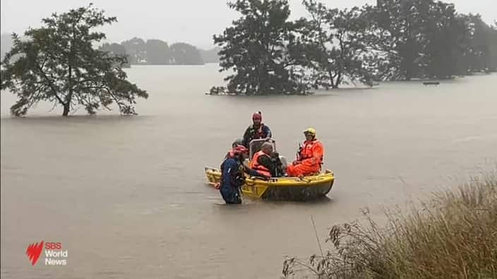 Three people are sitting in a yellow boat in brown floodwater, and one person is standing next to the boat.