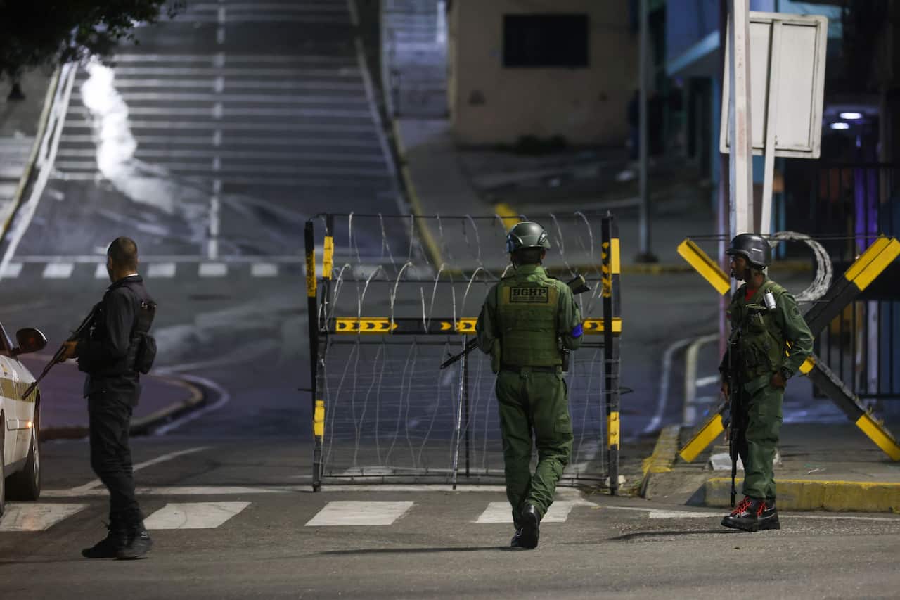 Three armed soldiers stand outside a compound with razor wire