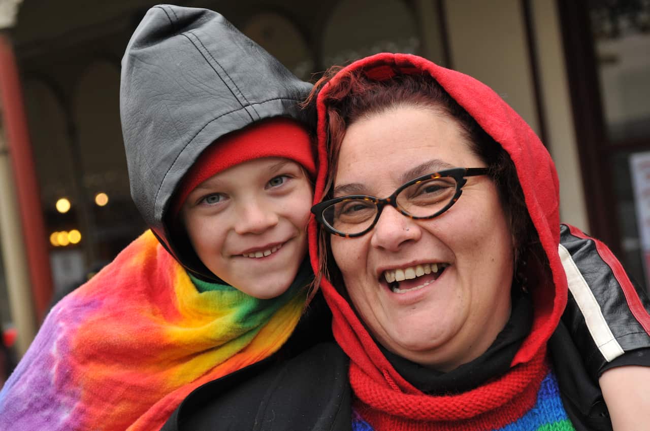 A child puts their arm around an adult. Both are smiling and wearing hoodies and are wrapped in a rainbow cloth