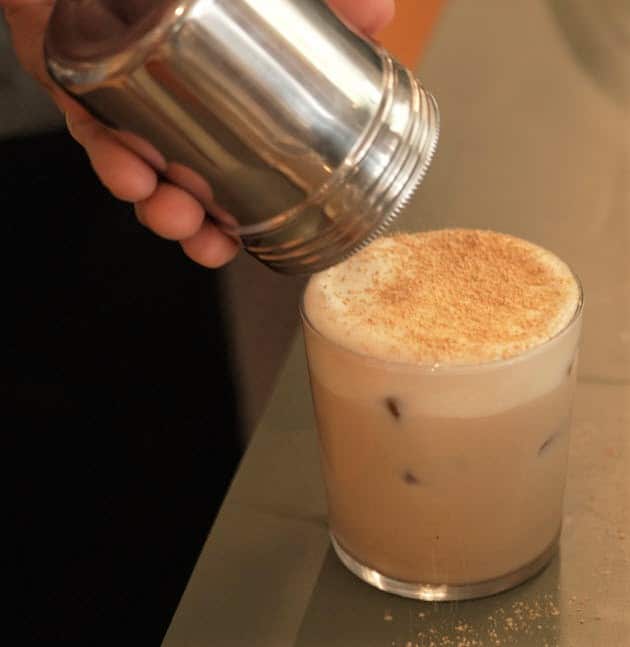 A frothy drink sits on a table, with chocolate powder being sprinkled on top.