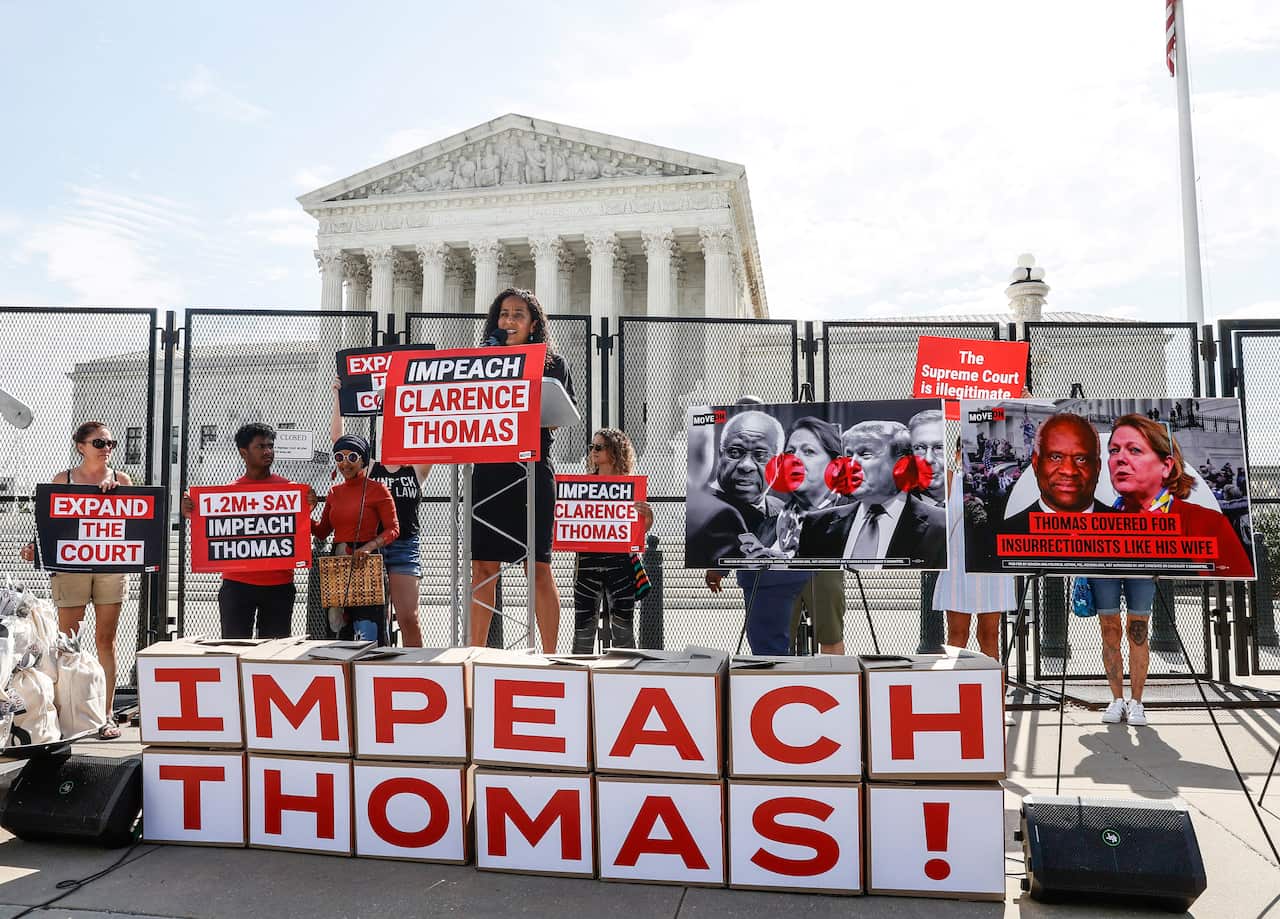 A woman dressed in all black delivers a speech at a podium in front of a sign reading, 'IMPEACH THOMAS!' in white and red block letters.