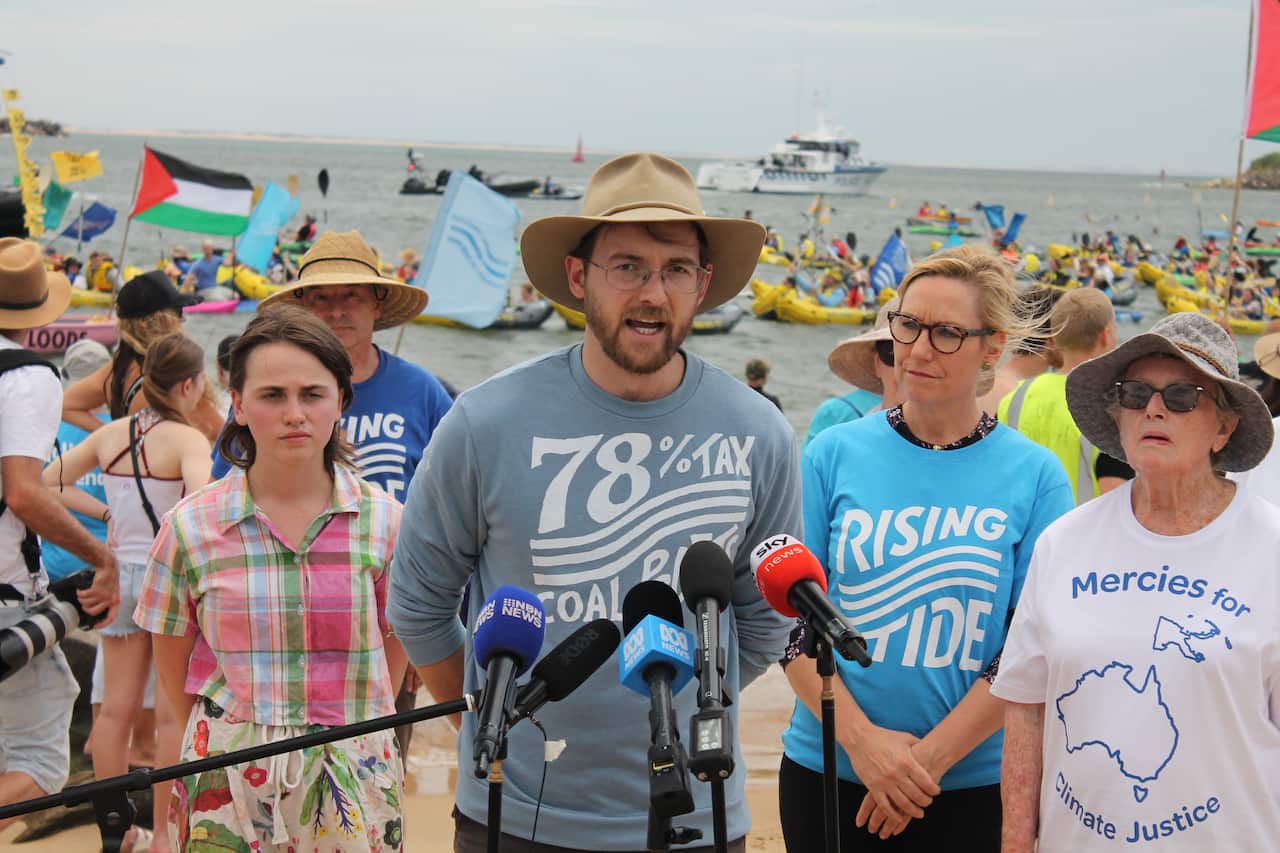 A man wearing a hat is flanked by three people as he speaks to the media outside. There is water behind him where a number of people on kayaks are paddling