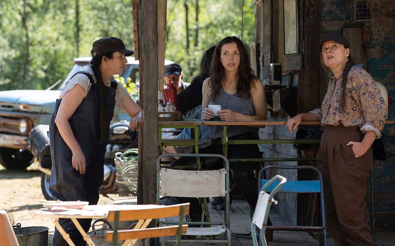 A woman sits at a tall bench on the porch of a rustic cafe. Two woman stand near her, one looking up at the roof, as if the three are mid-conversattion. 