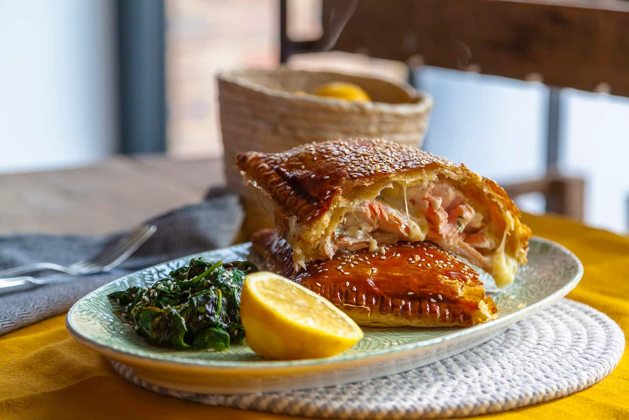 A gold-brown pastry parcel has been cut open to show a cheese and salmon filling. It sits on a green, oval plate along with cooked greens and half a lemon. 