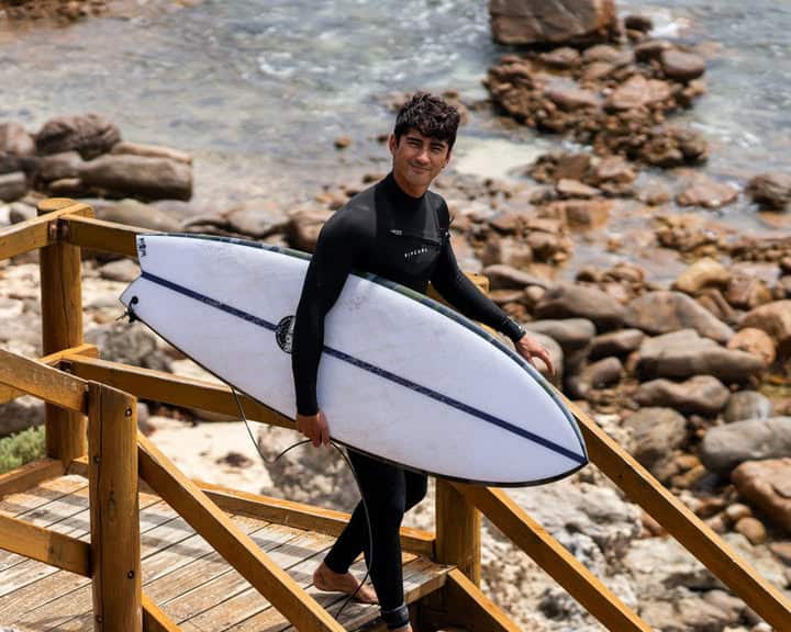 A man smiles as he walks down some wooden steps with a surfboard under his arm. There are small rocks and ocean water in the background