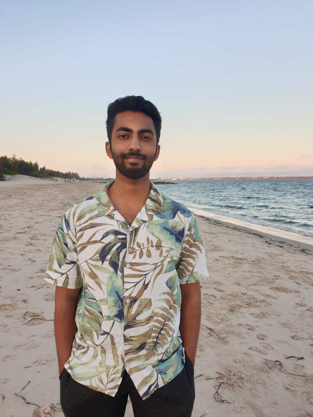 Sanjay stands on a beach wearing a short-sleeved button-up shirt with a leaf pattern.