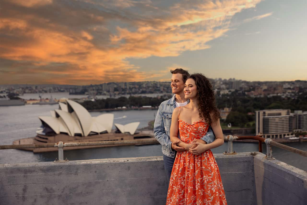 Nina Korbe & Billy Bourchier as their characters on top of the Sydney Harbour Bridge Pylon embracing each other. 