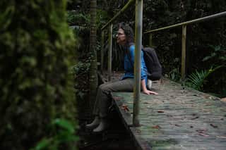 A woman in a blue shirt and grey trousers with a backpack is sitting on a bridge with her legs dangling, flanked by dense forest.