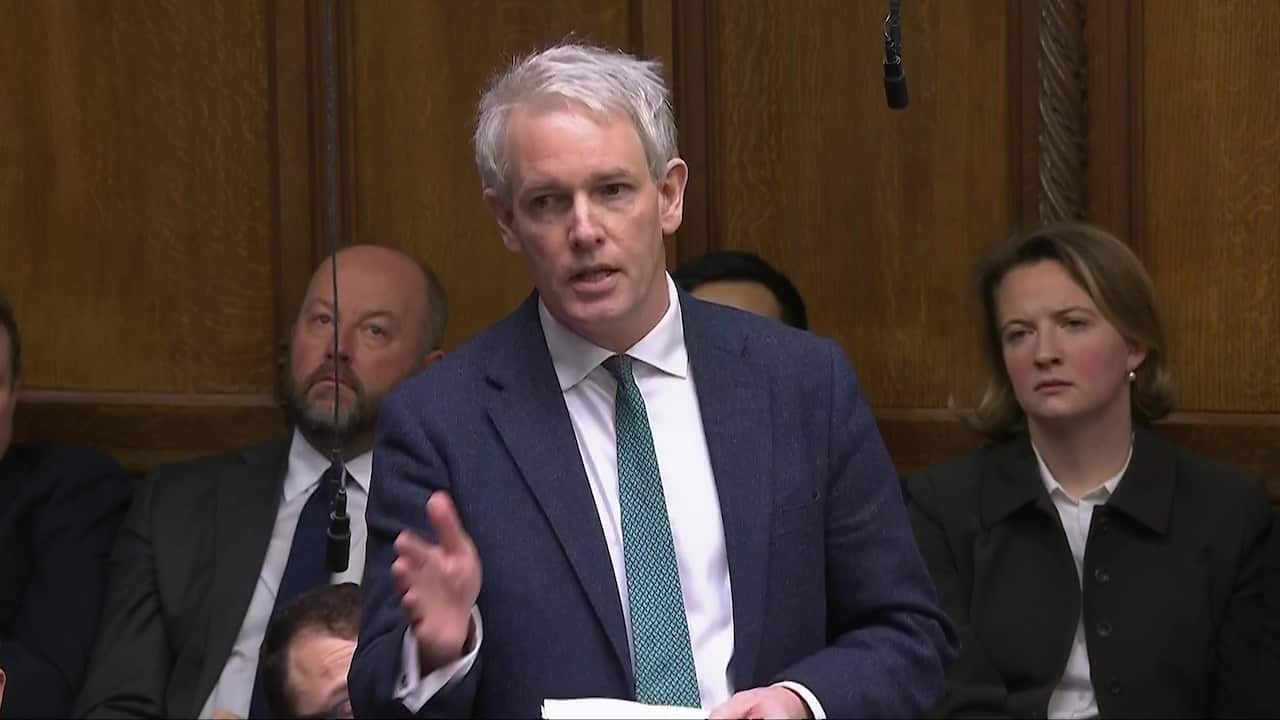 A man in a white shirt, teal tie and dark blue suit talking in parliament. People are sitting behind him.