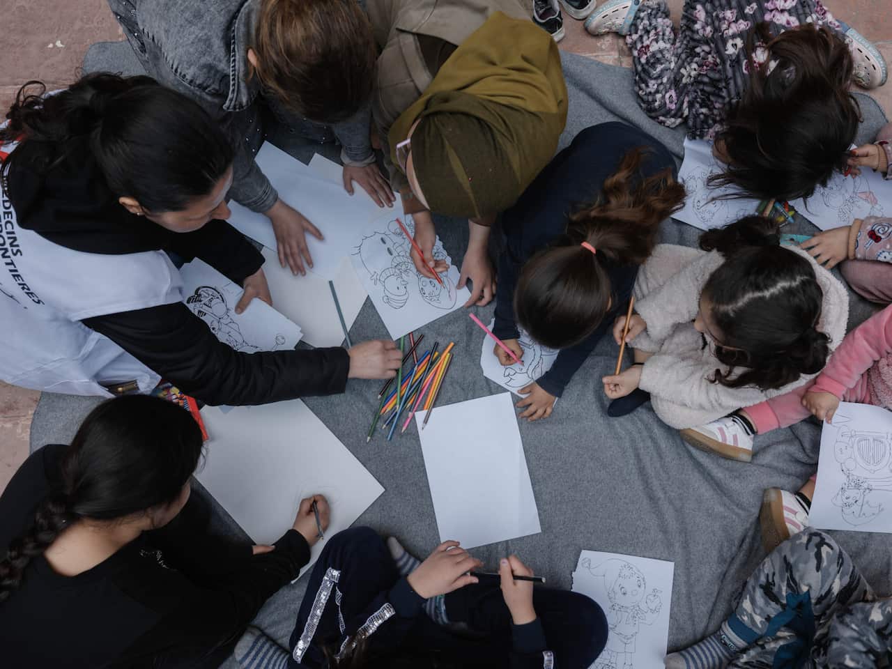 An aerial view shows women and children huddles over sheets of paper, some scribbling with pencils.